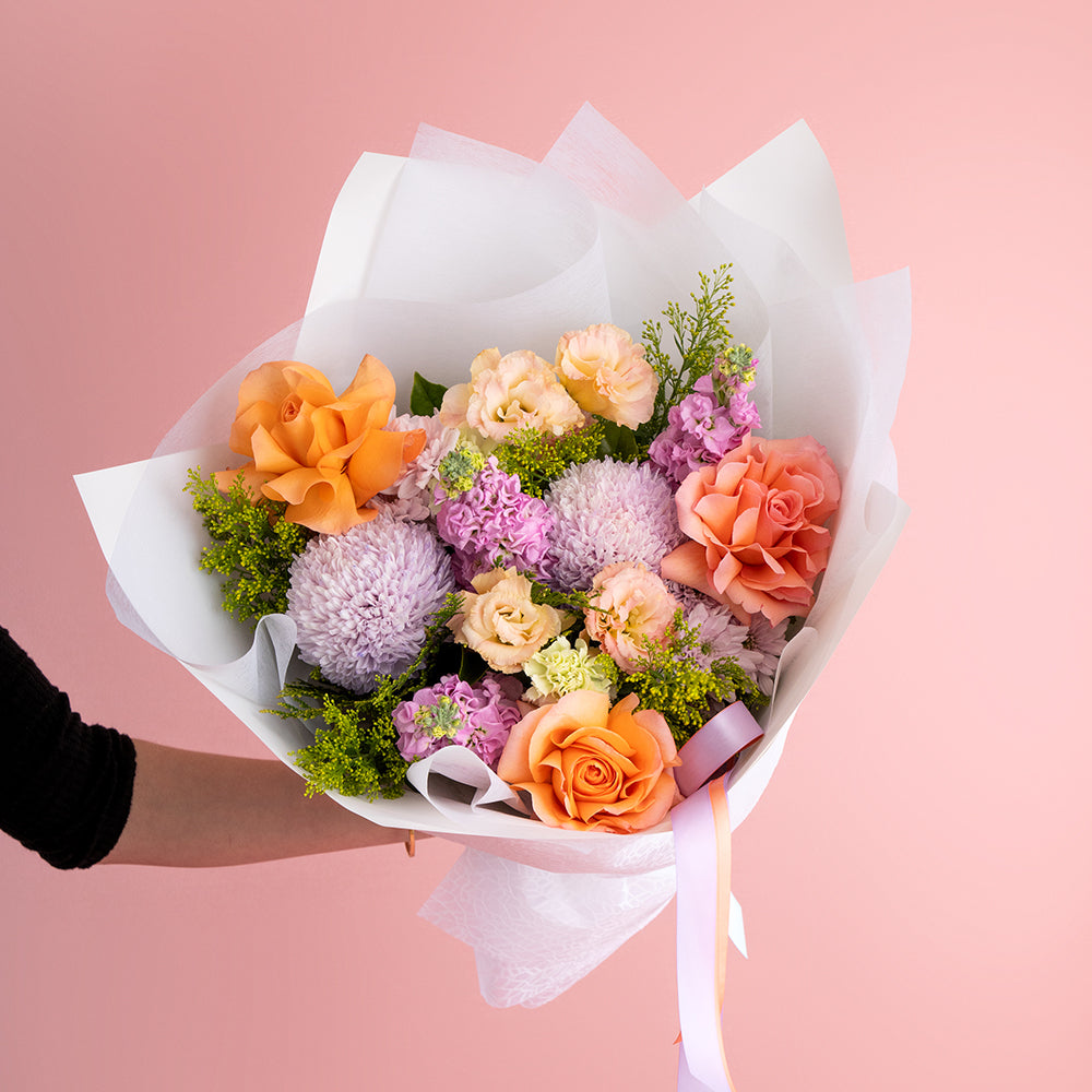 A bouquet of fresh flowers featuring shades of orange, peach, lilac, purple, pink, green, yellow, and other colors, arranged in a dynamic design with satin ribbon wrap.