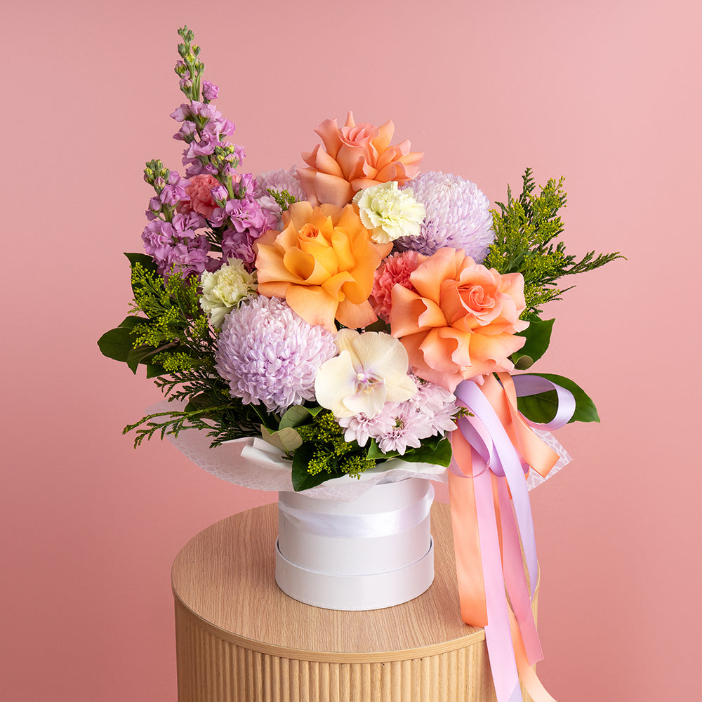 A vibrant floral arrangement featuring orange, peach, lilac, purple, pink, green, and yellow flowers, beautifully presented in a white hatbox with a pink ribbon.