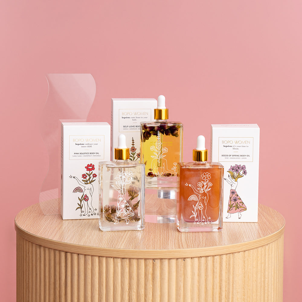 Three bottles of Bopo body oil in different colors with decorative packaging, displayed on a wooden surface against a pink background.