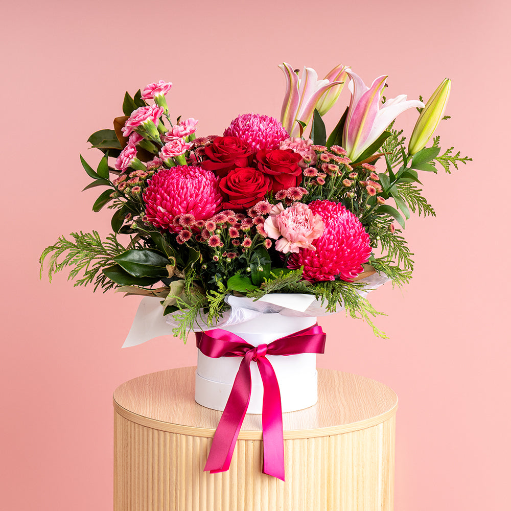 A floral arrangement with a mix of red, hot pink, soft pink, and purple flowers in a white hatbox, adorned with a pink ribbon.