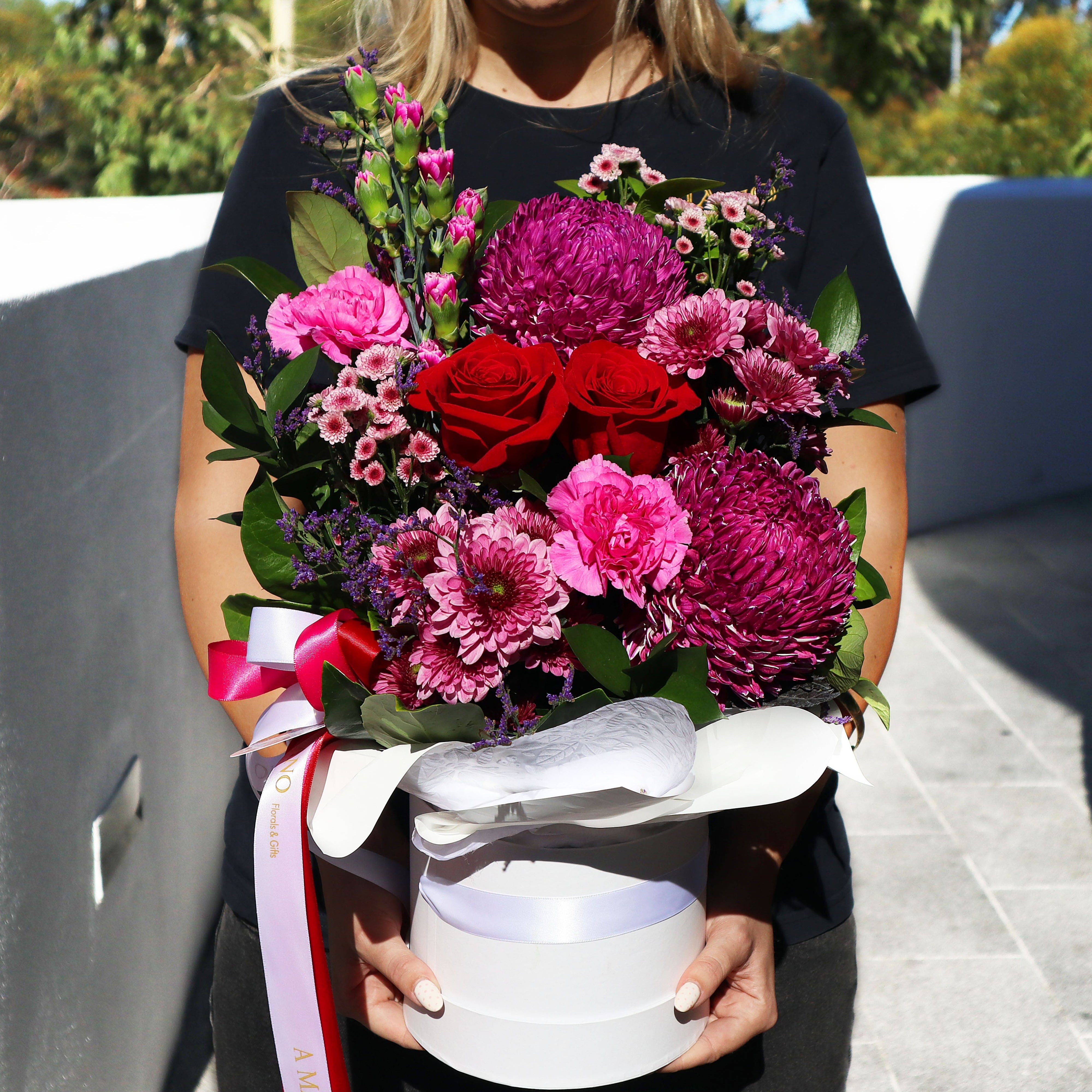 Person holding a bouquet of red and pink flowers in a white hatbox with white and red ribbon.