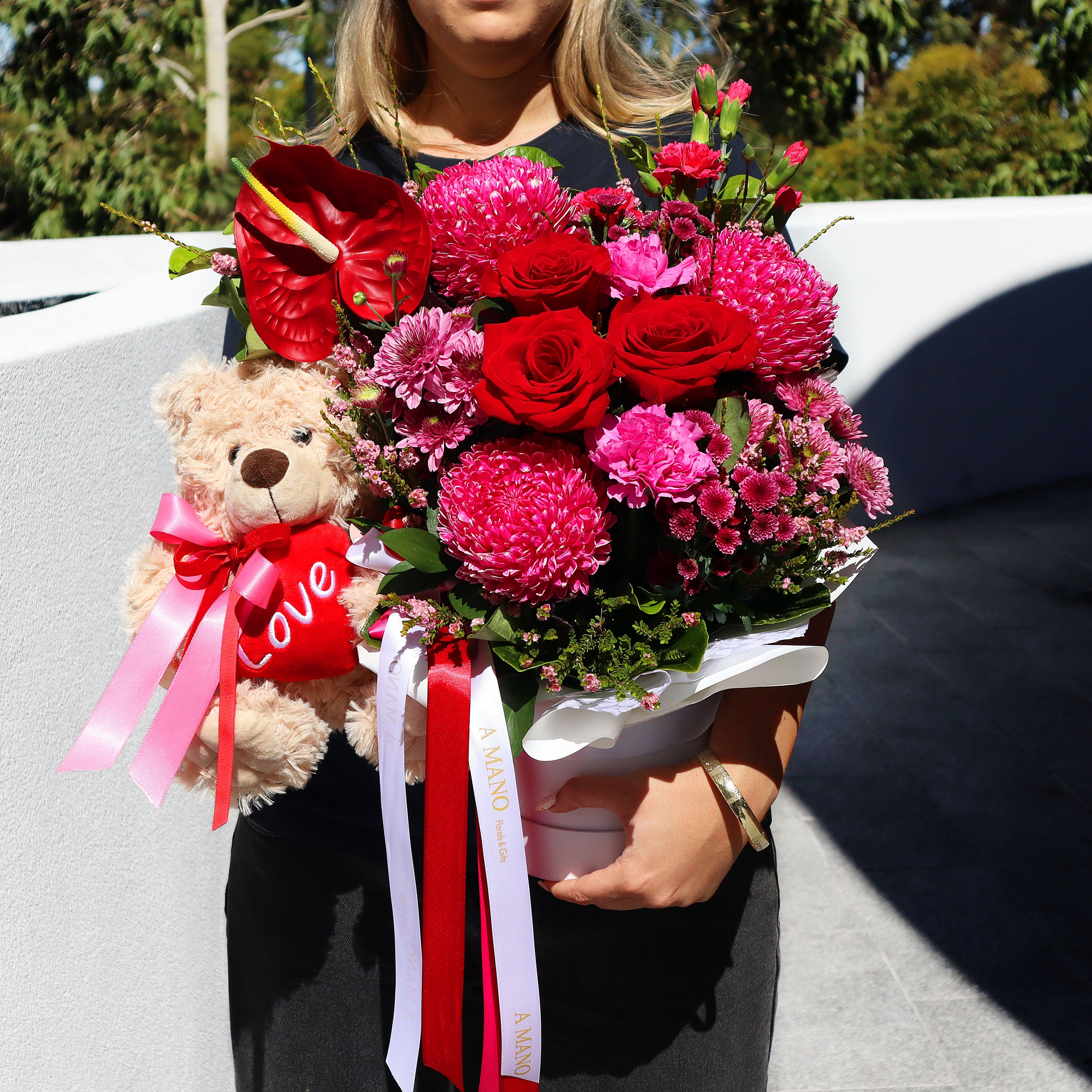 Person holding a bouquet of flowers and a teddy bear with a 'Love' heart accessory.