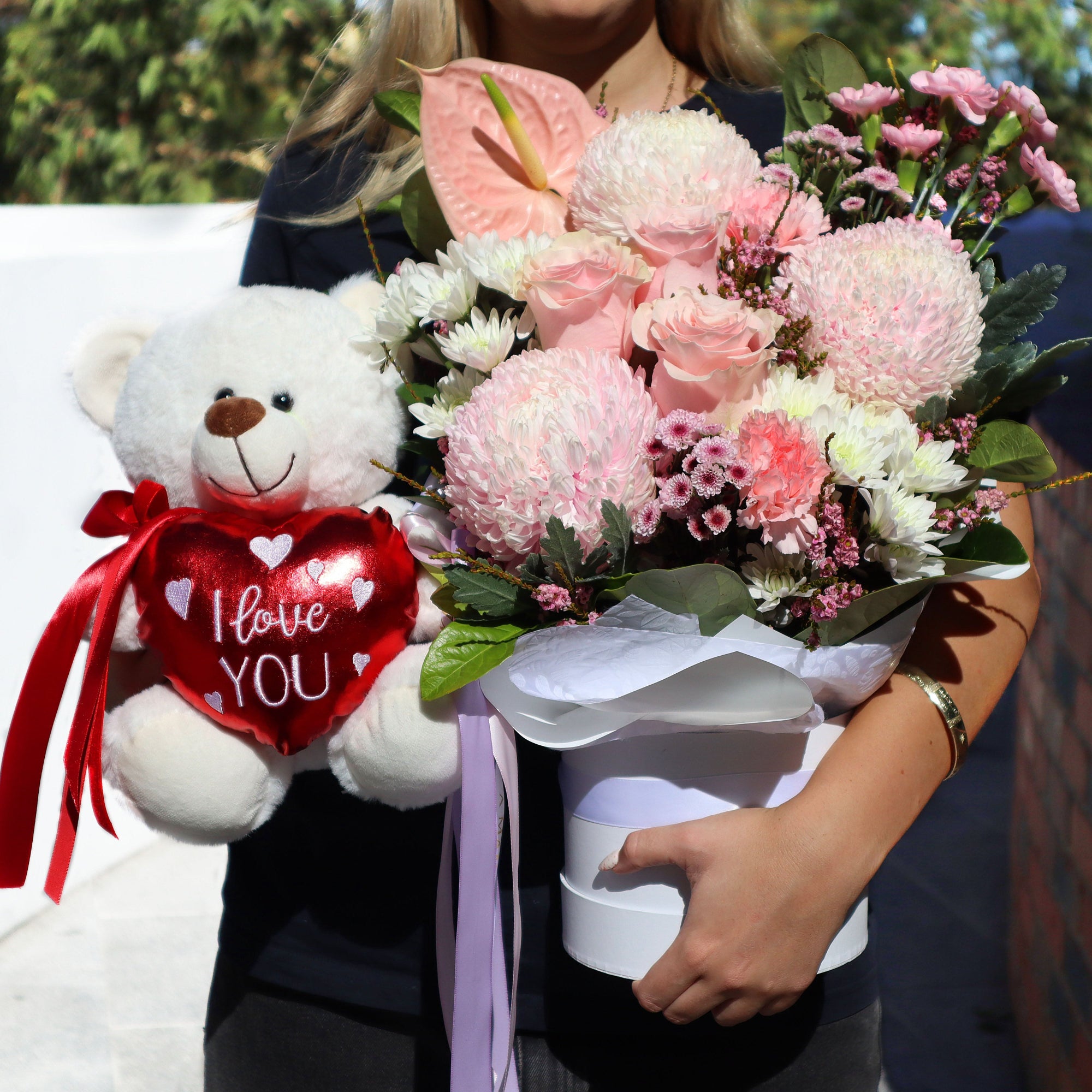 Person holding a bouquet of flowers and a teddy bear with a 'I Love You' heart.